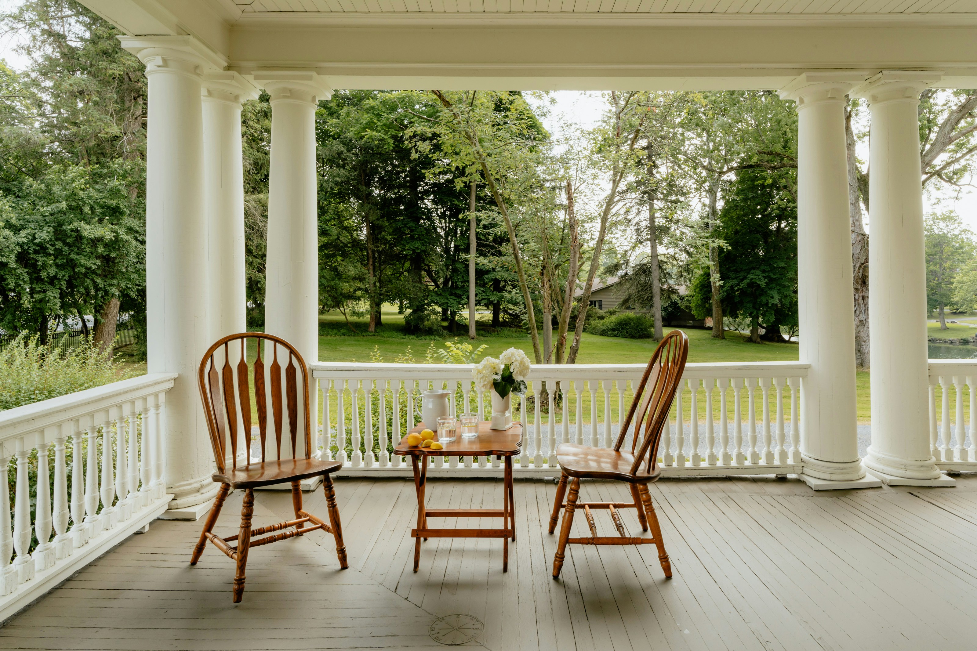 White classical porch with two wooden Windsor chairs, a folding table with a pitcher, lemons, and white hydrangeas, and a view of a green lawn
